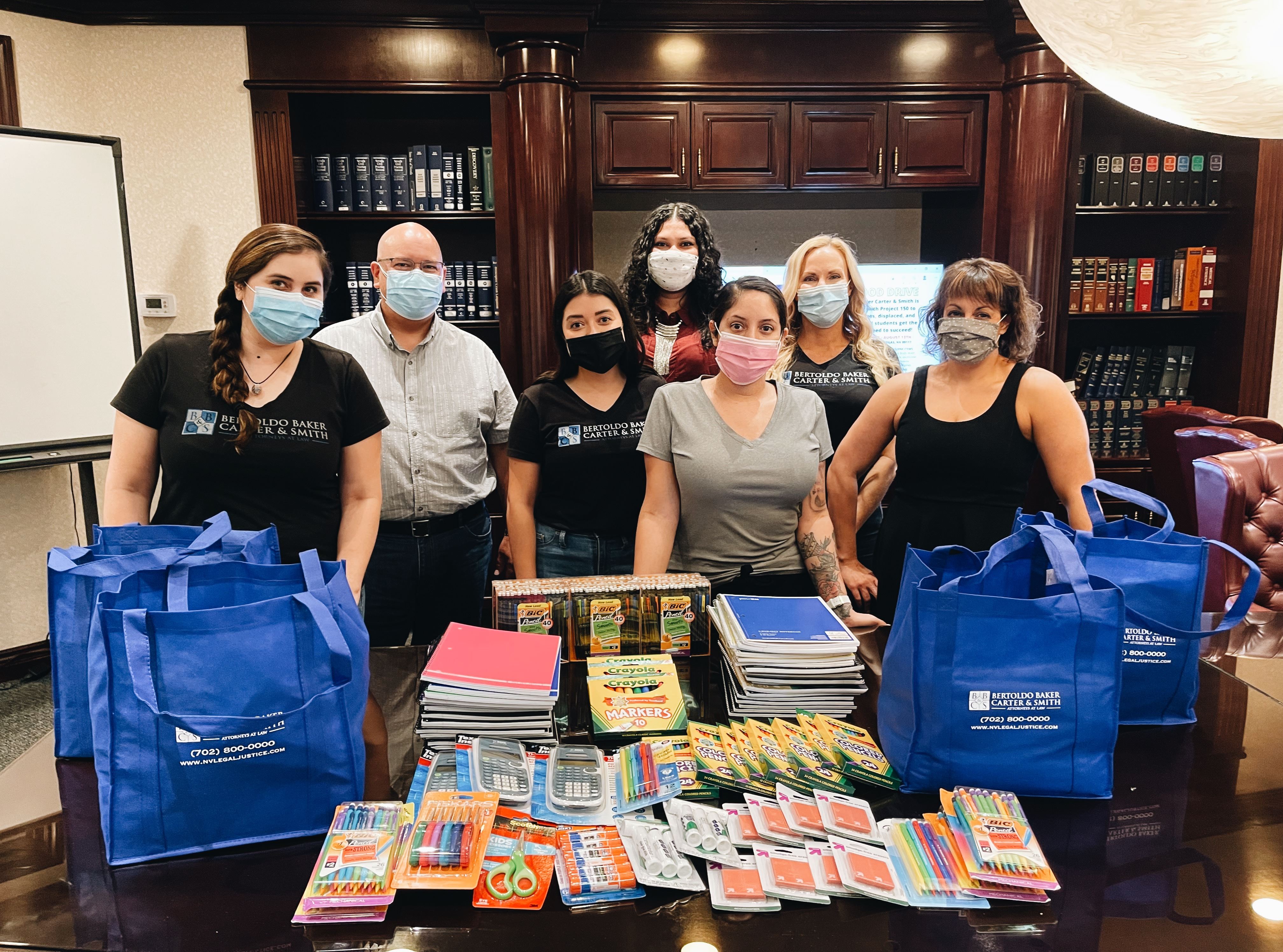 Picture of everyone at the firm who participated in the Local Back to School charity, standing in front of supplies such as paper, notebooks, markers, scissors, and glue sticks